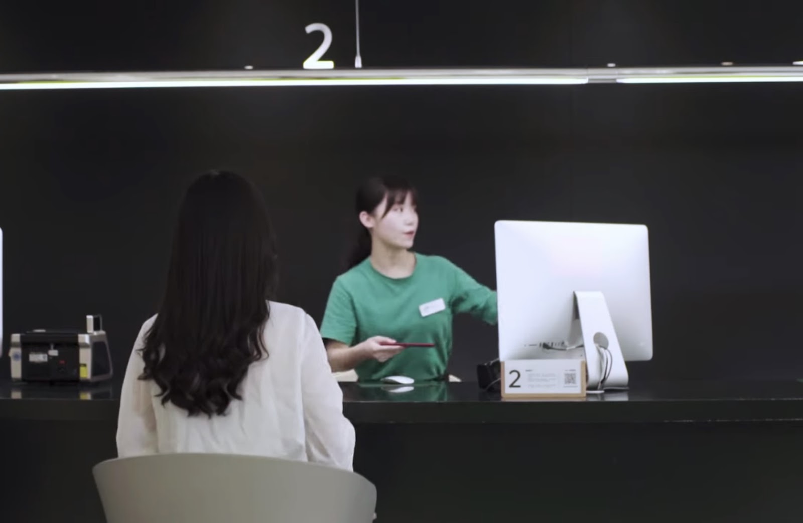 A customer service scenario where a female employee in a green shirt is assisting a customer with long dark hair seen from behind